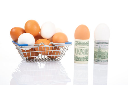 White and brown eggs in a metal basket next to rolled one- and two-dollar bills with an egg, set against a glossy white background for a financial food theme.の写真素材
