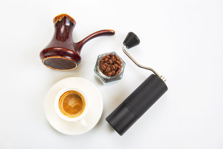 sleek black coffee grinder, a traditional ceramic cezve, a jar of coffee beans, and a white cup with fresh coffee, all on a clean white surface.の写真素材