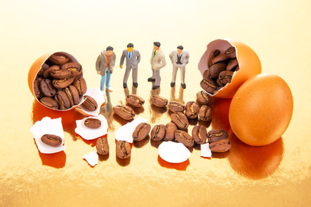Miniature figures in business and casual attire gather around cracked orange eggs spilling coffee beans on a golden surface, blending surrealism with coffee culture.の写真素材