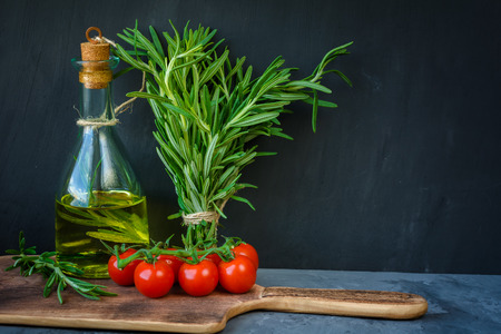 Fresh bunch of rosemary, cherry tomatoes, bottle of rosemary oil or olive oil on cutting board over stone table with copy space.の写真素材