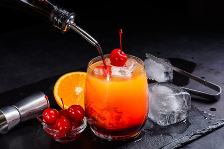 Tequila sunrise cocktail making. Grenadine slowly poured into a glass of orange juice, ice and tequila. Dark stone background with copy space.の写真素材