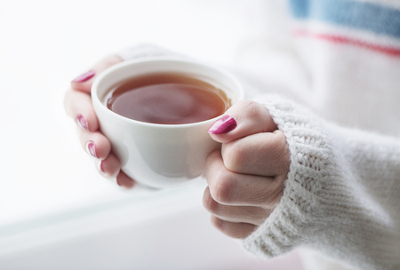 Tea drinking. The woman in a knitted, white pullover holds a cup of hot tea in her hands. Cozy weekends, winter drinks.の写真素材