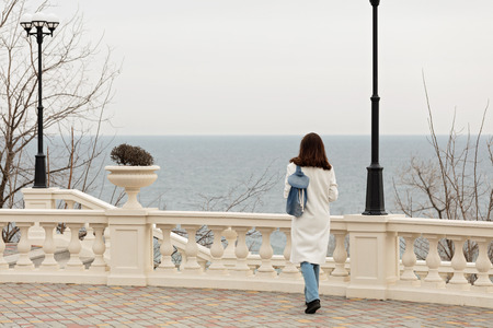 Young woman (brunette) in a white coat walks along the promenade. Selective focus.の写真素材
