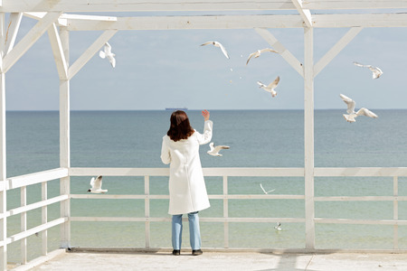Young woman (brunette) in a white coat feeds the gulls on the beach. Selective focus.の写真素材