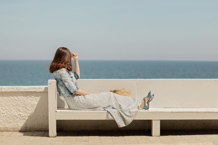 Young beautiful woman in a long skirt sits on a bench on a background of the sea horizon. Selective focus.の写真素材