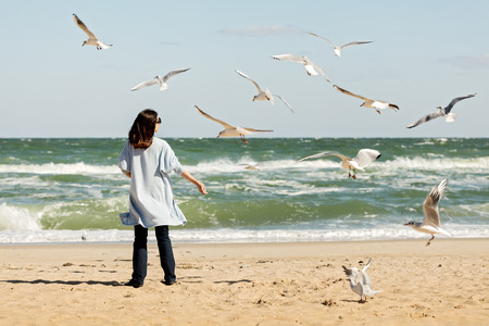Young woman (brunette) in a light blue cardigan and navy jeans, with a striped backpack, walks along the beach and feeds gulls. Selective focus.の写真素材