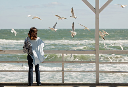 Young woman (brunette) in a light blue cardigan and navy jeans, with a striped backpack, walks along the beach and feeds gulls. Selective focus.の写真素材