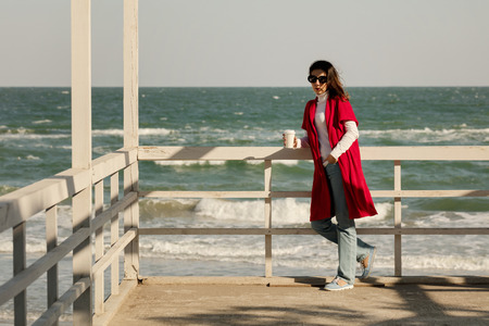 Young woman (brunette) in a red cardigan and light blue jeans walks along the beach and drinks coffee. Selective focus.の写真素材