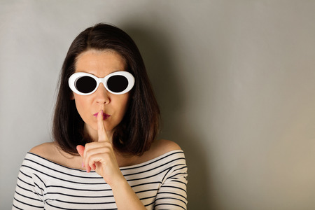 Woman in a white striped top and white sunglasses holds a finger near the lips. Selective focus.の写真素材
