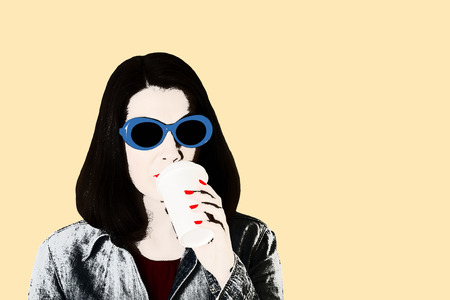 Photo in the style of pop art. Woman in denim shirt and blue sunglasses with red lipstick and nail polish drinks coffee. Isolated background.の写真素材