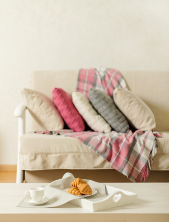 Beige sofa with plaid, colorful pillows (pink, grey, white) and coffee with croissantsin the living room. Selective focus.の写真素材