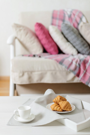 Beige sofa with plaid, colorful pillows (pink, grey, white) and coffee with croissantsin the living room. Selective focus.の写真素材