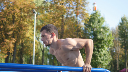 Athletic man doing push ups on parallel bars at sports ground in city park. Strong young muscular...