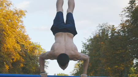 Strong muscular man doing a handstand in a park. Fit muscular male fitness guy doing stunts on...