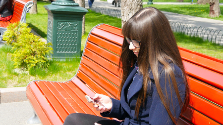 Young woman in glasses uses cell smartphone in a city park. Girl sitting on a red bench outdoor in spring and uses a mobile phone. Close-upの写真素材