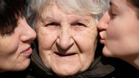 Young girl and adult woman kissing grandmother on cheeks, Granny smiling and looking to the camera. Three family generations. Grandmother, mother and granddaughter.の写真素材