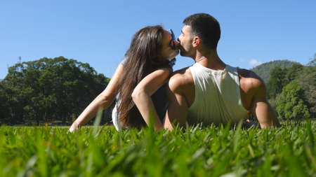 Young couple lying on green grass in park and relaxing. Man and woman sitting on meadow at nature and kissing. Girl and boy looking at the landscape and enjoying vacation. Rear Back Low angle of viewの写真素材