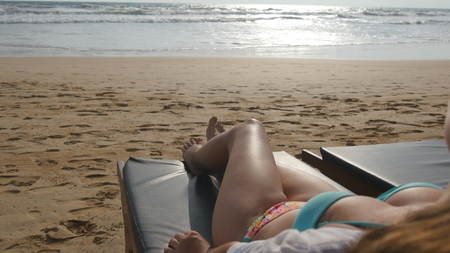 Female body on chaise-longue relaxing and enjoying during summer vacation on empty sandy ocean beach. Young woman lying on sunbed by the sea and tanning. Girl in bikini on a resort. Close upの写真素材