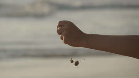 Female hand pouring sea sand through her fingers at sunset against an ocean background. Arm of young woman with sand strewed or falling from it. Grit drizzling from fist of girl. Close upの写真素材