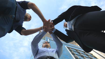 Three business men standing outdoor near office and stacked arm together in unity and teamwork. Hands of businessman team getting together in the center of a circle and then raised. Low angle of viewの写真素材