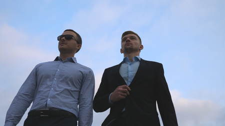 Two young businessmen walking in city with blue sky at background. Business men commuting to work together. Confident guys being on his way to office. Colleagues going outdoor. Slow motion Close upの写真素材