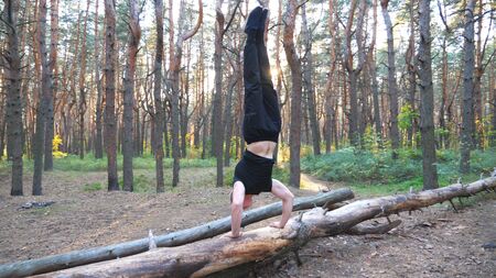 Strong man doing handstand on log in forest. Hardy and muscular guy doing stunts at wood. Athlete showing performance outdoor. Workout sport healthy lifestyle. Scenic background. Slow motionの写真素材