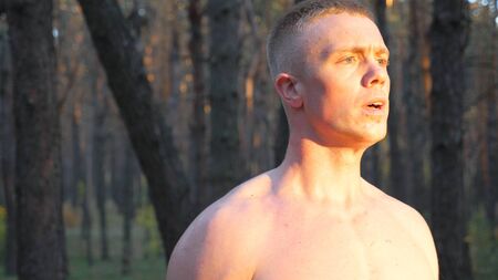 Muscular man with naked torso lifting hard weight training biceps. Strong guy working out in forest...