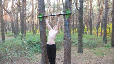 Strong and muscular man doing pull ups in beautiful forest. Male sportsman exercising on horizontal...