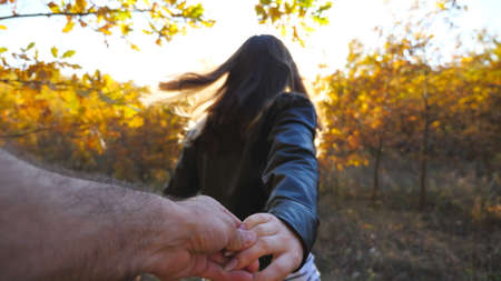Follow me shot of young woman pulls her boyfriend along trail through small oak trees. Happy girl holds male hand and jogs along path in garden. Beautiful nature at background. Concept of loving. POVの写真素材