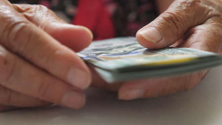 Elderly woman holds cash in arms and counts foreign currency over the table. Close up hands of old grandmother puts one hundred dollar banknotes on the desk. Money concept. Low view Slow motionの写真素材