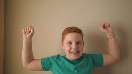 Happy male ginger child with freckles looking into camera and raises hands rejoicing achievement...