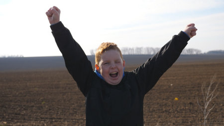 Little red-haired boy with freckles looks into camera and raises hands rejoicing achievement...