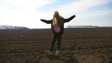 Cute female child with long blonde hair dances funny on plowed field at sunny day. Close up of happy...