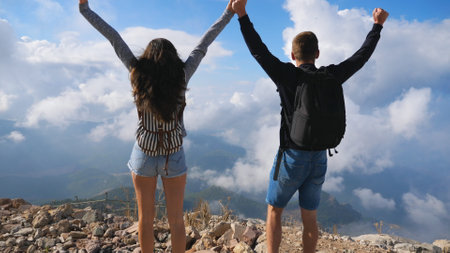 Couple of hikers with backpacks reaching up on mountain edge and raising his hands enjoying freedom. Man and woman actively resting during travel exploring new places together. Beautiful backgroundの写真素材