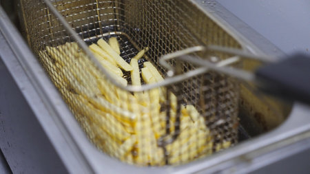 Male hand of cook shaking a lattice with french fries being prepared in hot boiling oil. Arm of chef lowering a grid with potatoes into deep fryer. Process of making fast food at kitchen. Top viewの写真素材