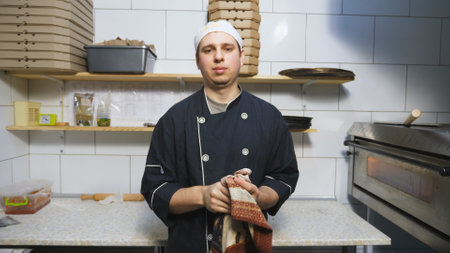 Portait of young cook in uniform standing at kitchen restaurant and wiping his hands with a towel after cooking dishes. Male chef looking at camera with a confident sight at cuisine cafe. Slow motionの写真素材
