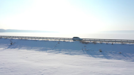 Aerial shot of white car riding through snow covered road near a frozen lake. SUV driving at countryside route near icy pond at sunny winter day. Flying over the auto moving through scenic landscapeの写真素材