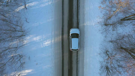 Aerial shot of white car riding through snow covered icy road. SUV driving at empty countryside route in the forest on winter day. Flying over the auto moving through scenic landscape way. Top viewの写真素材