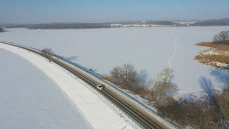 Aerial shot of car riding through snow covered road near frozen lake. White SUV driving at dam route on winter day. Flying over the auto moving through bridge of river. Scenic landscape way. Top viewの写真素材