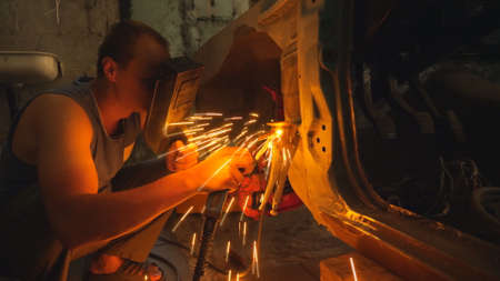 Repairer wearing protective mask welding some detail of auto. Mechanic working in garage or workshop. Man engaged servicing vehicles. Bright hot sparks flying around in dark. Car maintenance conceptの写真素材