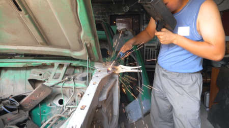 Professional repairer welding auto details using protective mask. Skillful mechanic fixing automobile. Man engaged servicing car in garage or workshop. Vehicle maintenance concept. Slow motionの写真素材