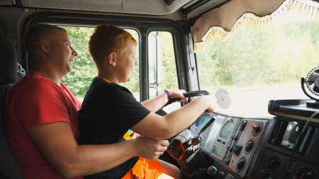 Little smiling red-haired boy holds the steering wheel sitting on lap of daddy and learning to operate the truck. Lorry driver teaches his ginger son to driving a car on country road. Dolly shotの写真素材