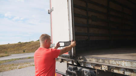 Male driver closing rear door of the truck at countryside. Lorry parked at country road at sunny...