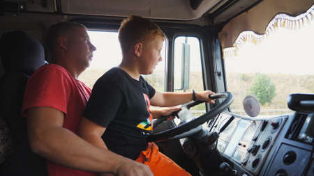 Father teaching his small red-haired kid to operate a car on empty country road. Focused little boy sitting on lap of dad and learning to drive the truck. Dad and son spending time together. Close upの写真素材