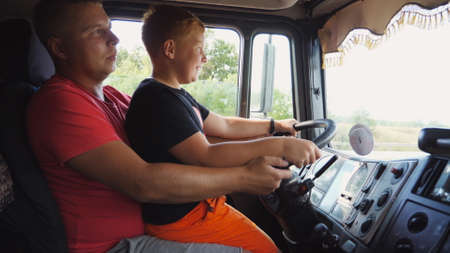 Little smiling red-haired child holds his hands on steering wheel sitting on lap of daddy and learning to drive the truck. Father teaches his ginger son to operating a car at country road. Close upの写真素材