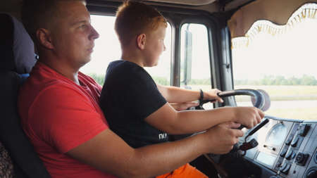 Little smiling red-haired child holds his hands on steering wheel sitting on lap of daddy and...