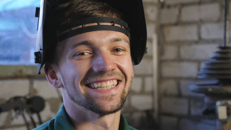 Close up of happy young welder opens protective mask after finishing his job. Portrait of mechanic in workwear smiling and looking into camera. Repairman with beard working in workshop. Dolly shotの写真素材