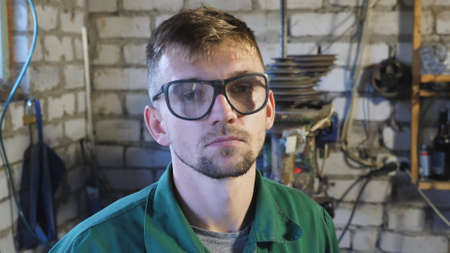 Professional mechanic in uniform putting on protective eyewear to get to work in workshop. Portrait of tired young repairman looking into camera at garage. Maintenance service concept. Close upの写真素材
