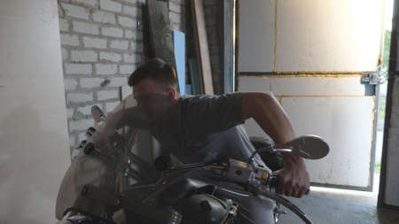 Young man pretending to fast ride a moto in workshop. Cheerful guy fooling around sitting on motorcycle in shed. Happy human enjoying and having fun in garage. Close up Slow motionの写真素材