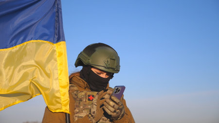 Female ukrainian army soldier using smartphone to read news or messaging with family. Girl in military uniform and helmet holding waving flag of Ukraine. Victory against russian aggression. Close upの写真素材